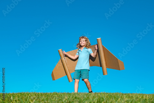 Child boy plays in an astronaut dreams of space. Happy child play with toy plane cardboard wings against blue sky. Kid having fun in summer field outdoor. Portrait of boy with paper wings.
