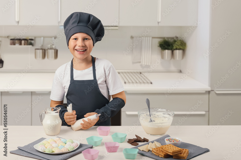 A Caucasian boy in a gray apron and cap holds a pastry syringe in his hand in a white kitchen and prepares cream, ice cream, dessert. Side view. A Caucasian boy in a gray apron and cap holds a pastry syringe in his hand in a white kitchen and prepares cream, ice cream, dessert. Side view.