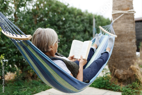 Smiling senior good-looking grey hair woman wearing glasses while reading in hammock in the summer garden