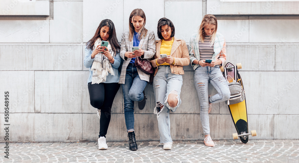 generation z multiethnic girls and friends using cellphone leaning against wall in university - obrazy, fototapety, plakaty 