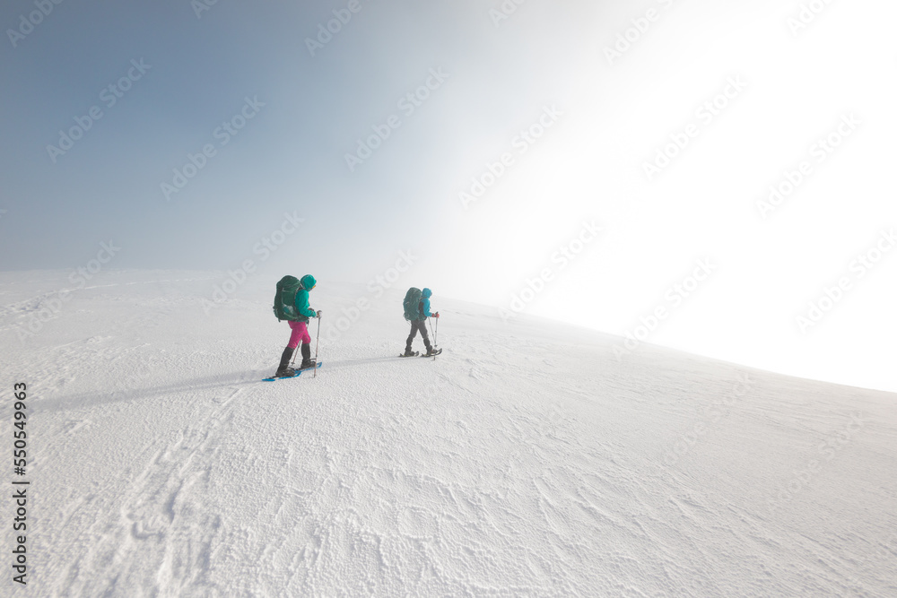 Two women on a winter hike. Girlfriends with trekking sticks go along a snow-covered mountain path. Girls with backpacks and snowshoes travel together.