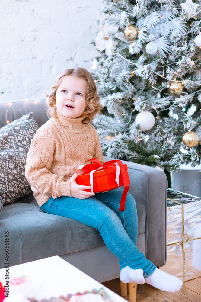 Naklejka premium cute happy little girl with curls is sitting on chair near Christmas tree with gift box in her hands, New Year and Christmas family concept