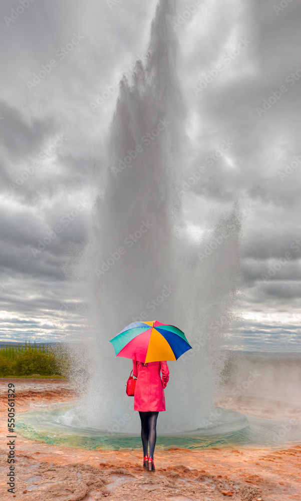 A beautiful woman in a red dress is standing with a multicolored umbrella watches Eruption of Strokkur Geyser in Haukadalur Valley - 
 Iceland