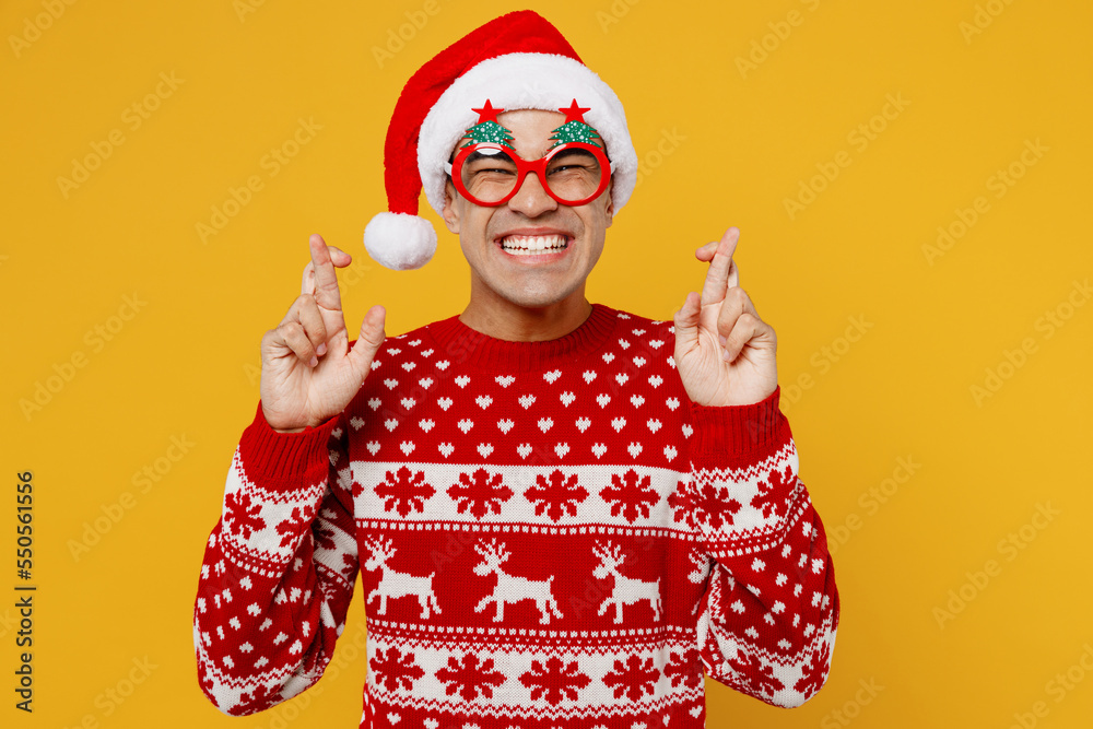 Merry young man wear red knitted christmas sweater Santa hat glasses posing wait special moment keep fingers crossed, making wish hope isolated on plain yellow background Happy New Year 2023 concept. Merry young man wear red knitted christmas sweater Santa hat glasses posing wait special moment keep fingers crossed, making wish hope isolated on plain yellow background Happy New Year 2023 concept.