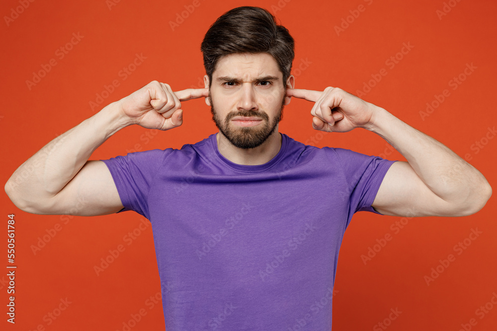 Young dissatisfied annoyed caucasian man wears casual basic purple t-shirt closed eyes cover ears with hands fingers do not want to listen scream isolated on plain orange background studio portrait. Young dissatisfied annoyed caucasian man wears casual basic purple t-shirt closed eyes cover ears with hands fingers do not want to listen scream isolated on plain orange background studio portrait.