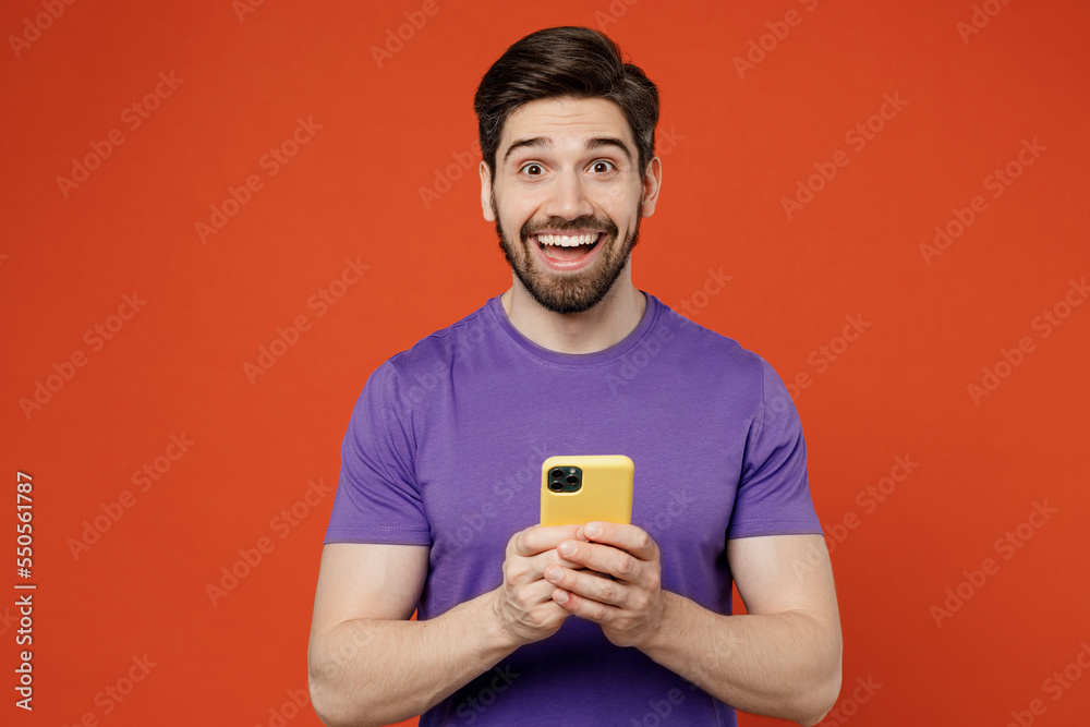 Young surprised brunet caucasian man wear casual basic purple t-shirt hold in hand use mobile cell phone in yellow case isolated on plain orange background studio portrait. People lifestyle concept. Young surprised brunet caucasian man wear casual basic purple t-shirt hold in hand use mobile cell phone in yellow case isolated on plain orange background studio portrait. People lifestyle concept.