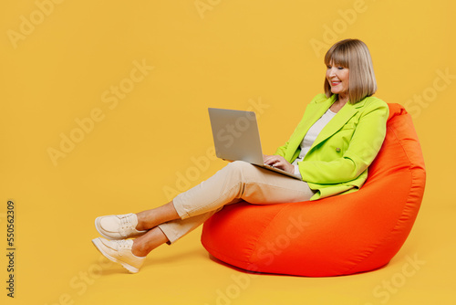 Full body elderly IT woman 50s years old wearing green jacket white t-shirt sit in bag chair hold use work on laptop pc computer isolated on plain yellow background studio. People lifestyle concept.