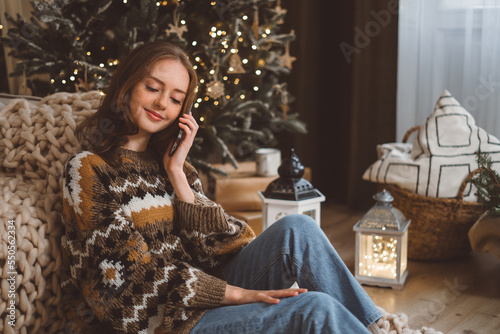 A charming young red-haired woman on the background of a Christmas tree in a cozy living room holds a mobile phone and waves her hand talks with her friend or family via video call