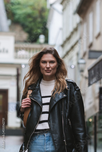 Young street fashion blond woman walking toward in urban city environment, serious face, determined,  looking at camera, black leather jacket and blue jeans. Waist up portrait crop.