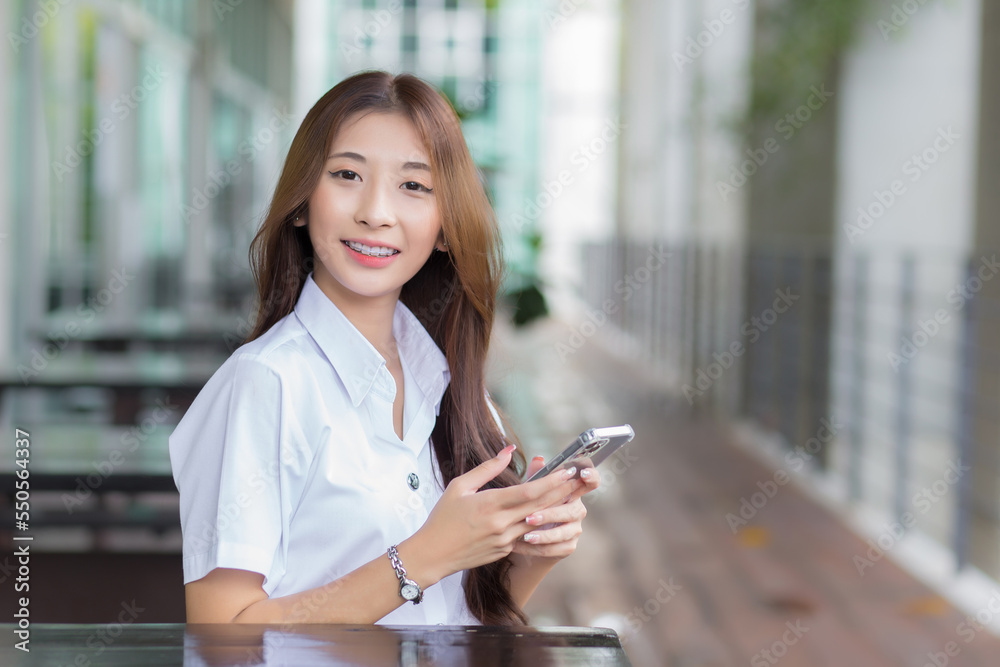 Portrait of an Asian Thai girl student in uniform is sitting work smiling happily and confidently while using smartphone in the building at university with background.