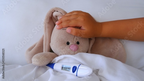 Child's little hand touches forehead of a sick toy bunny hare lying in bed with a high fever to check temperature with a thermometer. Medicine, healthcare and childhood concept