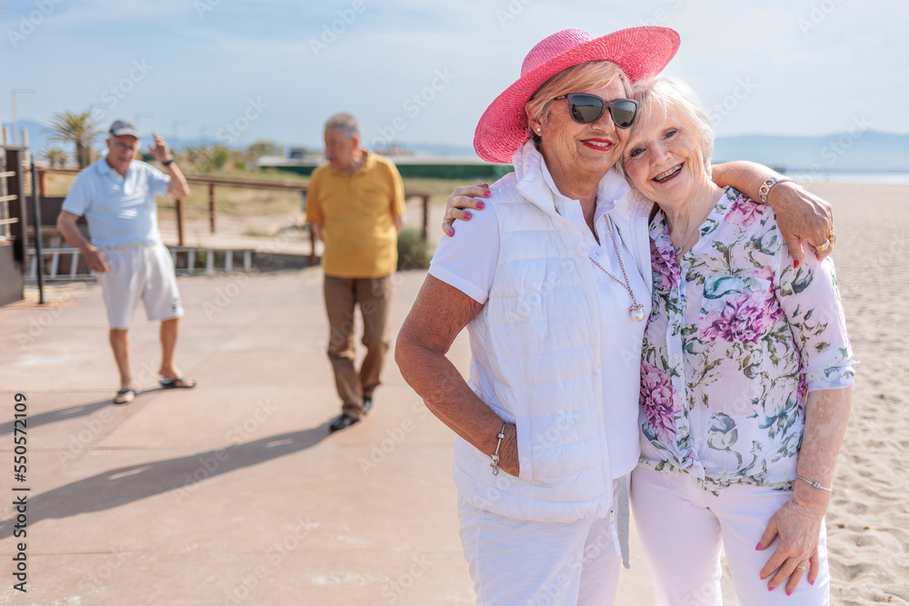 coppia di signore anziane si abbracciano affettuosamente mentre i mariti passeggiano sullo sfondo 