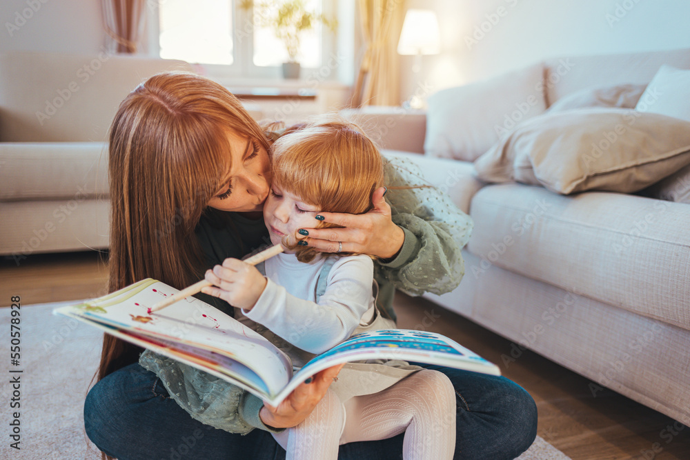 Attractive mother with little adorable daughter lying together at cushions on wooden warm floor in living room at home reading a book fairy tale close up. Weekends activities and development concept Attractive mother with little adorable daughter lying together at cushions on wooden warm floor in living room at home reading a book fairy tale close up. Weekends activities and development concept