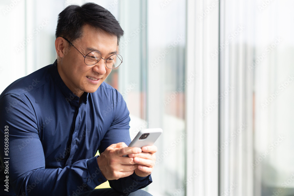 Closeup photo of a successful Asian businessman using the phone, a programmer in a shirt and glasses near a window inside an office messaging on social networks, using an application on a smartphone. Closeup photo of a successful Asian businessman using the phone, a programmer in a shirt and glasses near a window inside an office messaging on social networks, using an application on a smartphone.