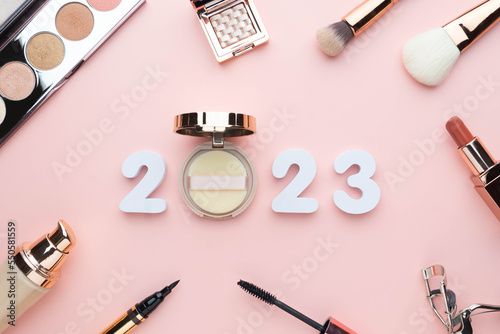 2023 Beauty cosmetic makeup products trends concept. Top view of 2023 white number with powder, lipstick, foundation, eyeshadow, brush, eyeliner and mascara on pink background.