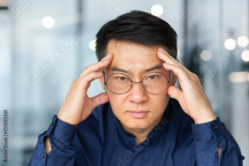 Close-up photo of thinking sad asian businessman, man in glasses looking at camera disappointed, boss in shirt inside office at work © Liubomir