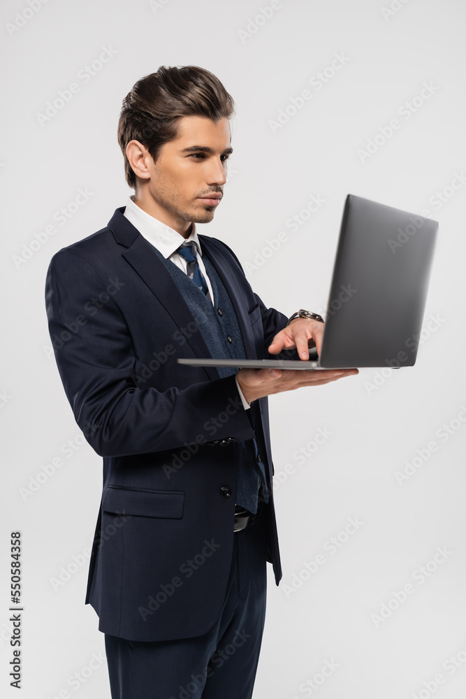 confident businessman in suit using laptop while working remotely isolated on grey confident businessman in suit using laptop while working remotely isolated on grey