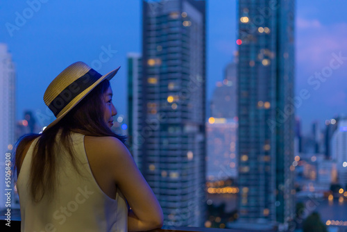 Asian woman tourist enjoying her urban city skyline of the nightlife view from the hotel room balcony for vacation and travel concept