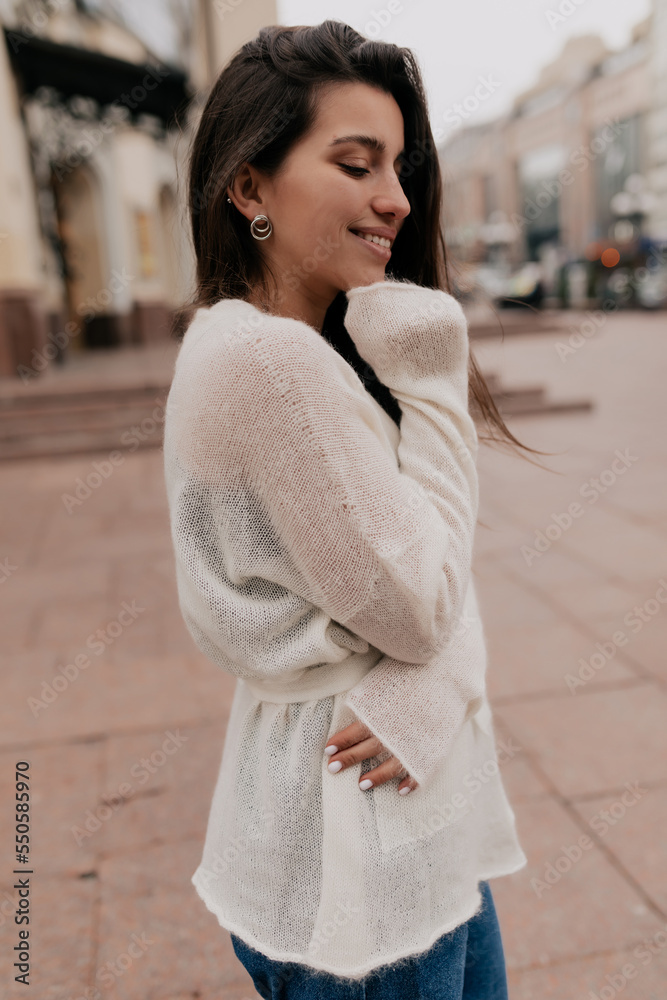 Romantic adorable smiling girl with long wavy hair and charming smile, looking down, touching her chin and walking in the city