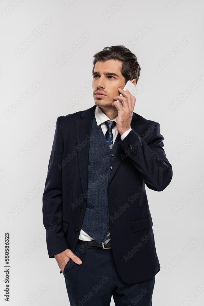 confident businessman in suit standing with hand in pocket and talking on smartphone isolated on grey confident businessman in suit standing with hand in pocket and talking on smartphone isolated on grey