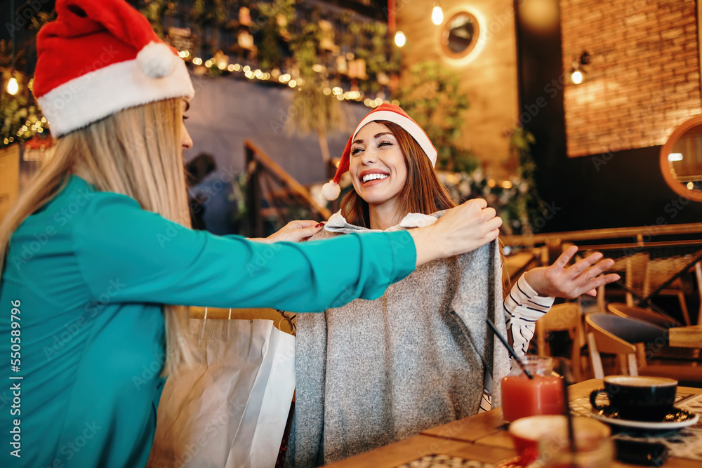 Two best friends with Santa hats sitting in coffee bar or restaurant after shopping and happily talking together. They also show each other the clothes they bought.