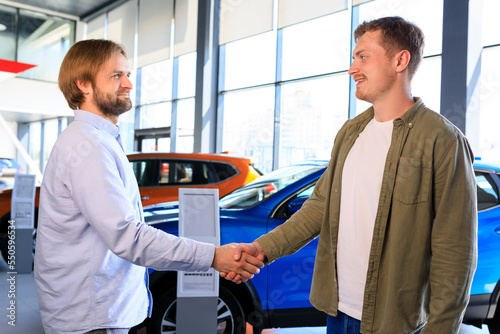 A sales manager shakes hands with a male customer on the background of a car dealership . Happy new car buyer