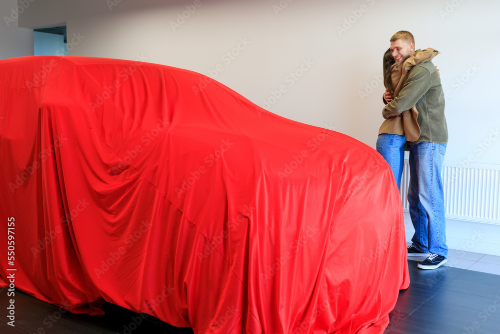 The man gave a car. A young woman embraces her man. There is a new car next to it under a red cloth