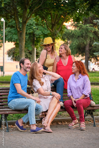 Portrait of five mature adults talking and laughing sitting on a bench in a park. © Ladanifer