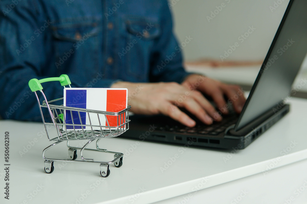 Small shopping cart with flag of France on the table. Man using laptop for shopping online.