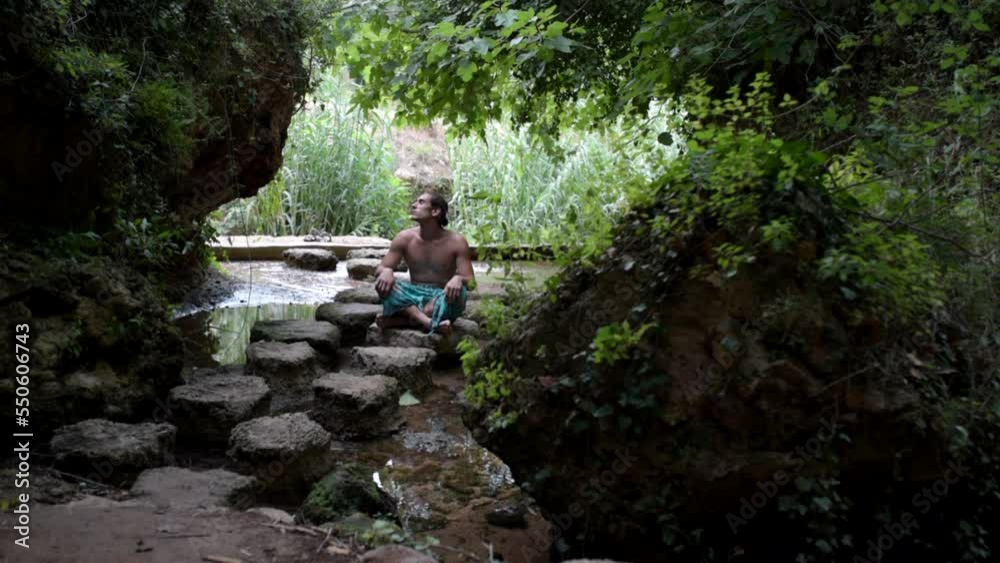 Caucasian man sitting on some stones by a small river Caucasian man sitting on some stones by a small river