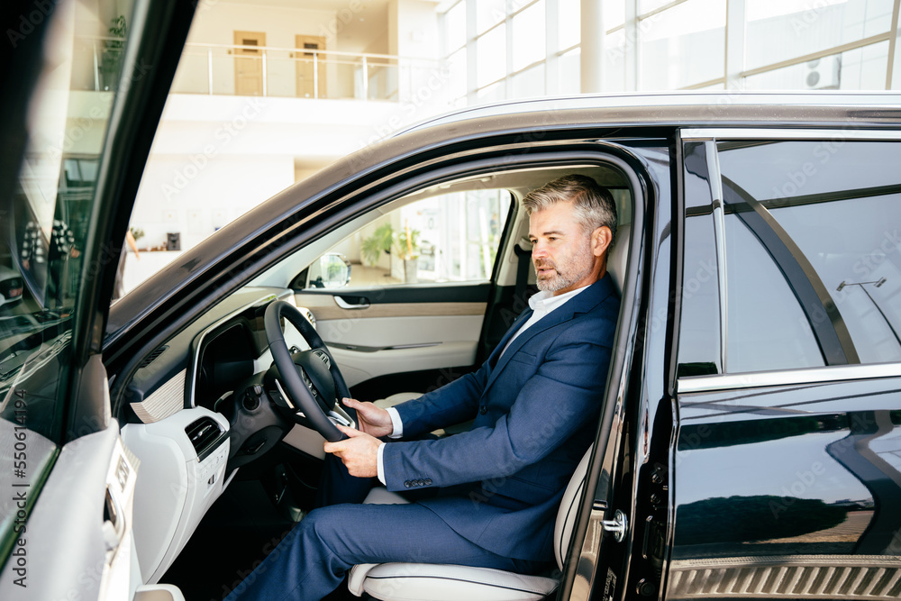 Side view of cheerful handsome senior senior man in formalwear sitting in car and looking the interior of his new auto in dealership.. Side view of cheerful handsome senior senior man in formalwear sitting in car and looking the interior of his new auto in dealership..