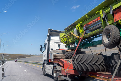 Heavy industrial truck with low side on low-frame platform transports disassembled big green combine harvester along ordinary highway on summer day.