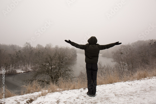 A man with outstretched arms stands on a hill near a river in winter