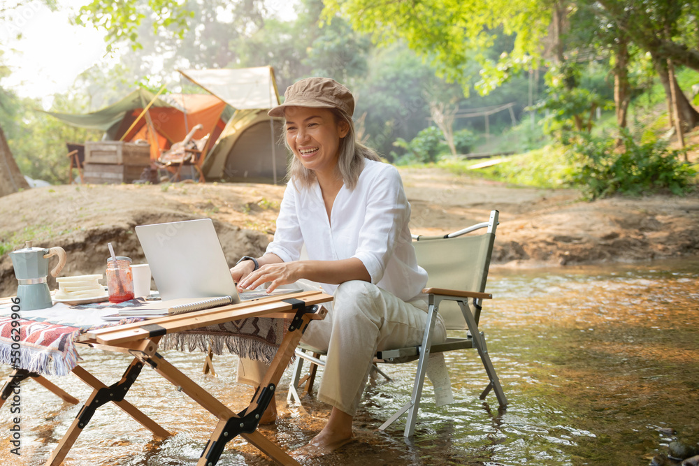 Fototapeta premium Asian woman travel and camping alone reading a book and using laptop while relaxing on the deck chairs in the river at natural park in Thailand. Recreation and journey outdoor activity lifestyle.