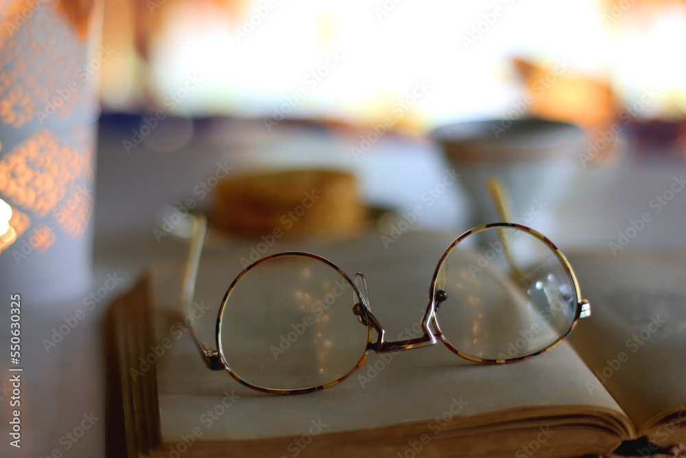 Decorative candle holder, cup of tea or coffee, plate of cookies, open book and reading glasses in front of the fireplace. Selective focus. Decorative candle holder, cup of tea or coffee, plate of cookies, open book and reading glasses in front of the fireplace. Selective focus.