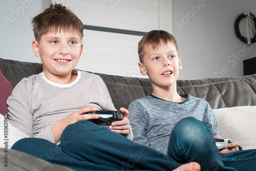Happy school aged boys sit on sofa with game joysticks. Children play videogames. Interesting amusement, competition.