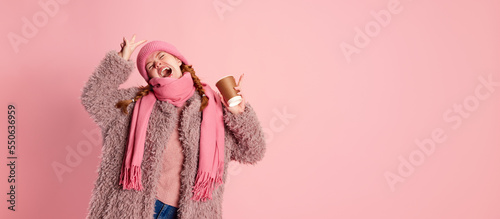 Portrait of emotive young girl in knitted hat, scarf and furry coat, posing with cup isolated over pink background. Hot winter drinks