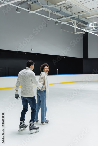 Cheerful african american woman holding hand of boyfriend while ice skating on rink