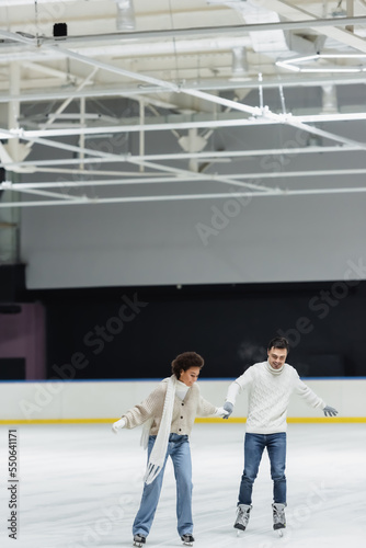 Smiling multiethnic couple in gloves and sweaters ice skating on rink