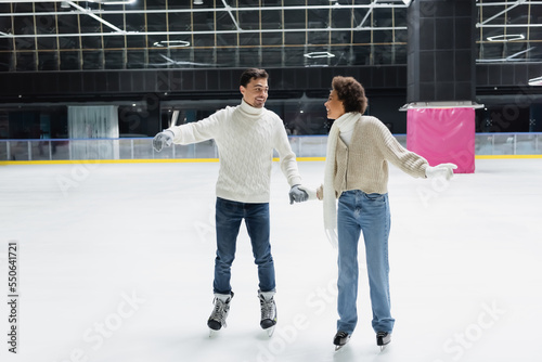 Smiling interracial couple in gloves and sweaters holding hands on ice rink