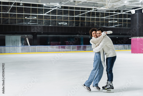 Multiethnic couple in warm clothes embracing and looking at camera on ice rink