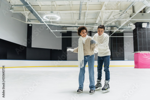 Cheerful man hugging and teaching african american girlfriend on ice rink