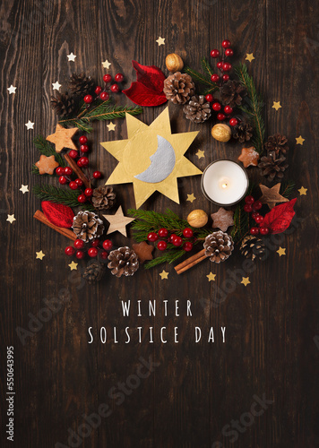 Winter solstice day, December 21. Sun and moon symbol, Christmas trees, pine cones, paddub branches with red berries, candle on dark wooden background, top view. photo