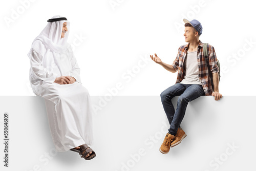 Male student sitting on a white panel and talking to a mature arab man wearing ethnic clothes