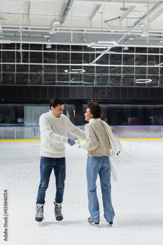 Positive multiethnic couple holding hands while skating on ice rink during date
