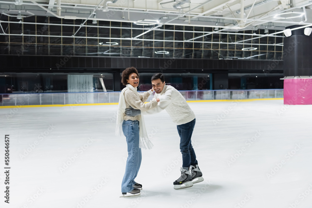 Positive multiethnic couple in gloves looking at camera while having fun on ice rink