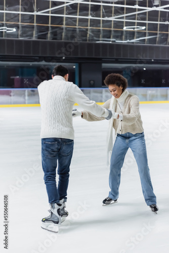 Smiling african american woman holding hands of boyfriend in ice skates on rink