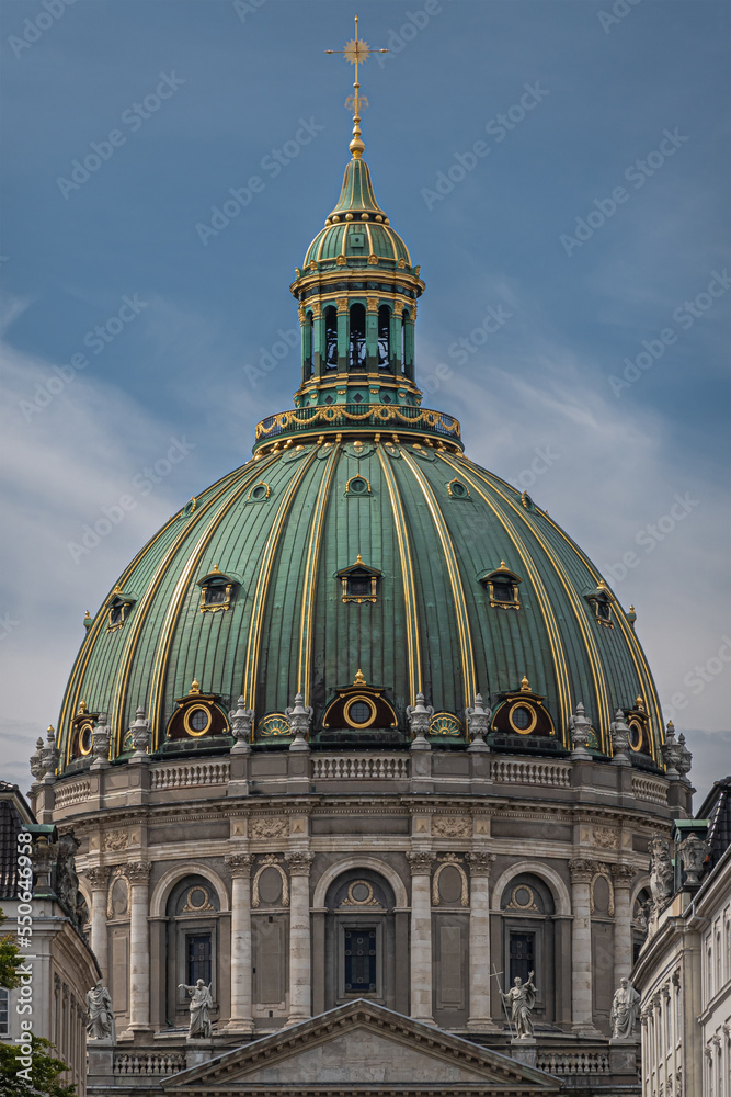 Copenhagen, Denmark - July 24, 2022: Closeup of green dome with golden cross pinnacle and trims above gray stone circular wall with statues and pediment of Frederik's church under blue sky