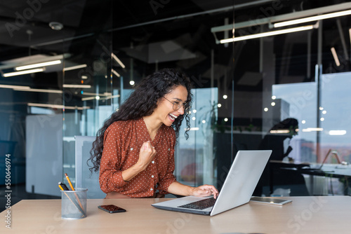 Hispanic businesswoman celebrating victory and successful achievement, office worker received online news and happy smiling and looking at laptop screen holding hand up triumph gesture.