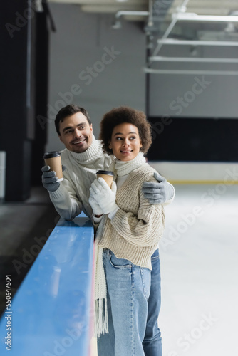 Smiling man hugging african american girlfriend with coffee to go on ice rink